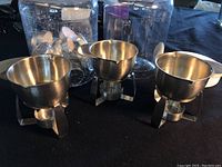 Photo of three stainless steel butter warmers with candle holders, arranged on black cloth background.