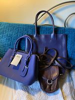 Three purple purses placed on a blue quilted surface with a metallic bedframe in the background, showing overall condition and style.