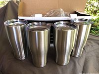 Four stainless steel 30 oz tumblers side by side with clear lids placed on a brown cloth surface with open box in background.