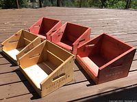 All five wooden storage containers arranged in two rows on a wooden deck, showing red and blonde finishes with angled fronts and hand cut-outs.