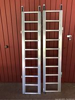 Pair of aluminum bifold ramps standing vertically against a red wall, showing front side with ladder rungs and folded hinge points.