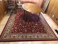 Full view of the rectangular red area rug under a dining table with a floral pattern and beige bordered edges.