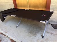 Full view of assembled cot showing black fabric surface, aluminum frame and legs with rubber feet on concrete floor.