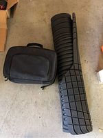 Photo showing black soft-sided emergency kit case and rolled flexible auto cargo mat on concrete floor.