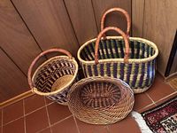 Photo showing four of the baskets grouped on a tile floor against a wood-paneled corner wall. Includes large rectangular basket with handles and multiple smaller baskets inside and nearby.