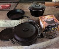 Full view of 3 cast iron Dutch ovens, one boxed (new), fry pan and flat skillet, showing coil handles and surfaces.