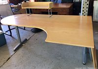 Main desk showing light wood grain laminate top, curved cutout edge, and two adjustable metal legs. Shelf with raised edges on metal supports attached at rear of desk.