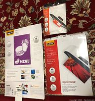 Photo showing two packs of Fellowes laminating pouches and one package of luggage tags on floral carpet background.