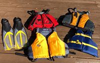 Photo showing full lot including three paddling jackets, one youth life jacket, and one pair of yellow and grey Cressi Pluma swim fins size 7-8.