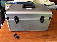 Closed silver metallic lock box with black handle and two keys on wooden table.