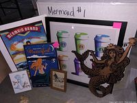 Photo showing multiple mermaid-themed framed prints, posters and a wooden mermaid cutout figure grouped together, illustrating the lot's varied items.