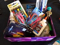 Box of office and school supplies with glue, scissors, pens, pencils, page markers, and rubber bands visible