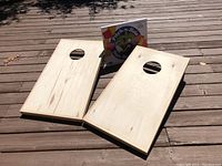 Two light-colored wooden cornhole boards with circular holes, laid flat on a deck surface, no paint or markings.