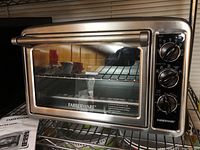 Front angle view of Farberware stainless steel toaster oven, showing glass door, wire rack inside, and three black control dials on right side.