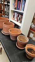 Five terracotta clay pots arranged in a line on a black surface, with bookshelves and furniture in the background