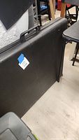 View of the black folding table leaning against a chair, showing the handle and overall rectangular shape.
