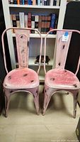 Two pink metal chairs with distressed finish side by side on floor in front of bookshelf