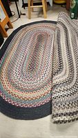 Two oval braided rugs shown side by side, one partially folded back to show the second rug beneath.