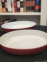 Two oval red ceramic casserole dishes stacked one inside the other showcasing the white interior and red exterior with books in the background.
