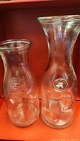 Two clear vintage glass milk bottles with embossed logos placed side by side on orange surface.