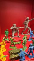 Multiple plastic toy soldiers and accessories displayed on a red background, showing various poses and colors including a tank and horse figure.