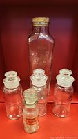 Five vintage glass storage bottles displayed against a red background, showing size and stopper variety