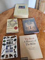 Photo showing all five books on a wooden table: Holy Bible hardcover, 'The Jesus Dynasty', 'The Tomb of Christ', 'Understand the Bible', and 'The Book: A History of The Bible'.