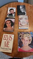 Photo of six hardcover biography books on a wooden table, showing front dust jackets featuring portraits of Lady Diana, Elizabeth Taylor, Brooke Shields, Goldie Hawn, Paul Reiser, and Elvis Presley.