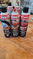 Photo showing three front aerosol cans of FastWax branded car care products on wooden surface. Labels visible with product names including TS2 Tire Shine, FW1 Cleaning Wax.