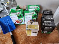 Lot of Christmas decor items placed on a wooden table including four boxes of Christmas lights, three boxes of black light clips, one compact blue snow shovel with black handle, and one boxed exterior light fixture.