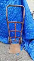 Front view of heavy duty metal dolly leaning against a blue tarp, showing flat platform and handle with horizontal supports.