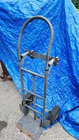 Front view of the metal hand dolly standing upright with curved handle and platform base on ground, against blue tarp.