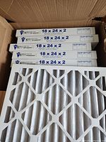 Box with 5 visibly stacked new furnace filters and a filter laying atop displaying white pleated filtration material with cardboard frame and diamond grid pattern.