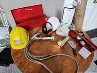 Overhead photo showing both hard hats, kitchen faucet parts, red toolbox, first aid kit, and various tools arranged on a round wooden table.