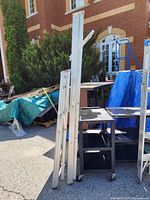 Two aluminum ladders standing upright outdoors with wood and tarps in the background.