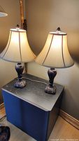 Two 27-inch table lamps with beige fabric shades on a black side table.