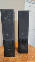 Front view of two Missil floor standing speakers with fabric covers and visible dust, placed on wooden surface.