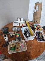 Wide view of the office supply items arranged on a wooden table including two adding machines, baskets with pens and pencils, stapler, tape rolls, and other small supplies.