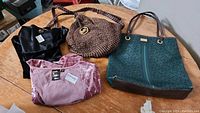 Four items on wooden table: one green Tommy Hilfiger tote bag, one brown woven Michael Kors handbag, two tops (one black, one soft pink) with tags visible