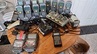 Photo showing multiple handheld retail scanners aligned behind a table with charging docks, label printers, and various cables.