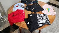 Round table with all clothing items arranged including black Dean T-shirts with white graphics, Fender dress shirts, infant onesie, and two large ball caps.