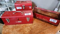 Three red metal tool boxes on a wooden surface, two with latches and handles, one open-top heavily worn box with visible rust and a size sticker.