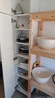 Photo showing upper cabinet shelving with stacked metals pots, pans, and mixing bowls alongside wooden shelving with large white ceramic mixing bowls.