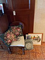 Full view of wooden chair with floral cross-stitched seat and two pillows on it, three framed art pieces leaning against the wall nearby.
