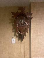 Front view of the wooden cuckoo clock hanging on a beige textured wall, showing the carved leaves and bird figure with pinecone weights and Roman numeral clock face.