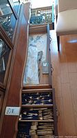 Drawer open showing a wooden tray with compartments containing various silver-tone flatware and napkin rings on a blue felt liner.