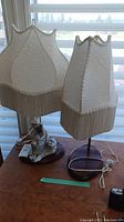 Pair of lamps on wooden table, one with Lladro figurine base and fabric shades with beige fringe