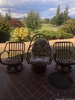Three wicker/rattan outdoor chairs positioned on a tiled porch, two without cushions and one with a cushioned seat and backrest, overlooking a garden view.