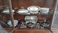 Photo showing multiple silver plated serving dishes and trays inside a glass cabinet on shelves with reflective surfaces.