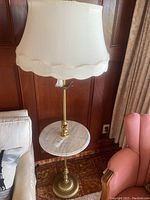 Full view of the standing floor lamp showing the gold-toned base, circular marble table, ornate cherub figures, and cream fabric lampshade.